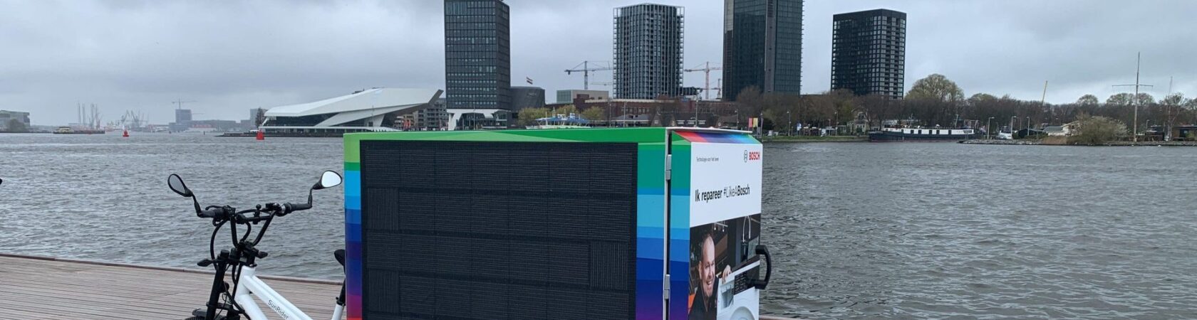 Een elektrische bakfiets met Bosch-reclame staat geparkeerd aan de waterkant met een skyline op de achtergrond.