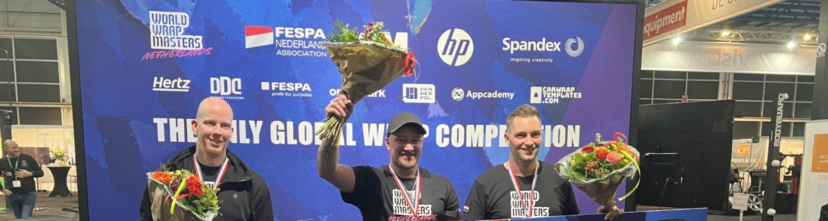 Drie mannen poseren trots met hun prijzen en bloemen op een podium tijdens de World Wrap Masters competitie.