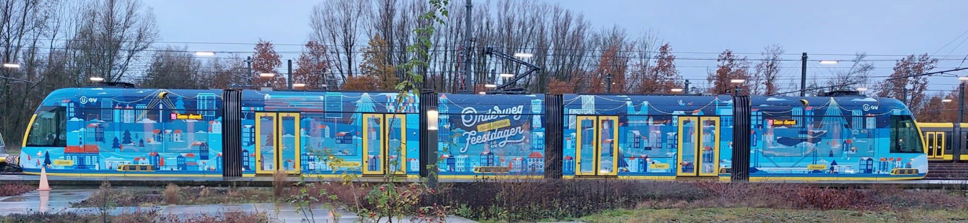 Een kleurrijke tram met een feestelijke winterdecoratie staat op het spoor omgeven door bomen en herfstbladeren.