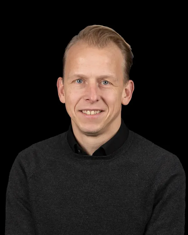 Een glimlachende man met een donkere trui tegen een zwarte achtergrond.