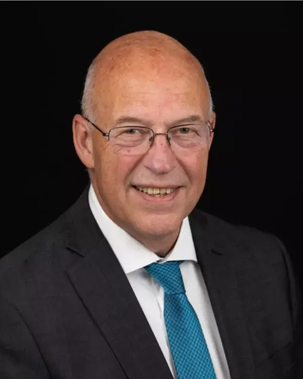 Een man met een blauwgrijze das en een donker pak poseert voor een zwarte achtergrond.