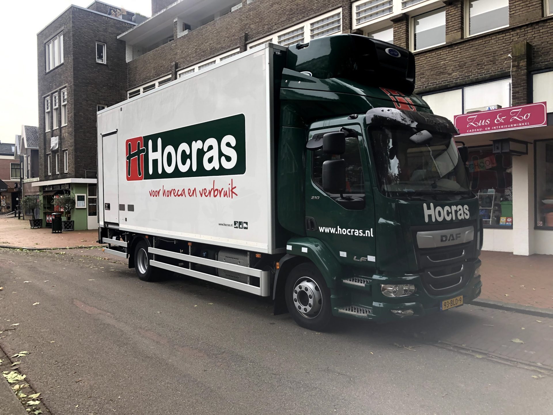 Een vrachtwagen van Hocras geparkeerd naast een gebouw en een winkel met de naam 