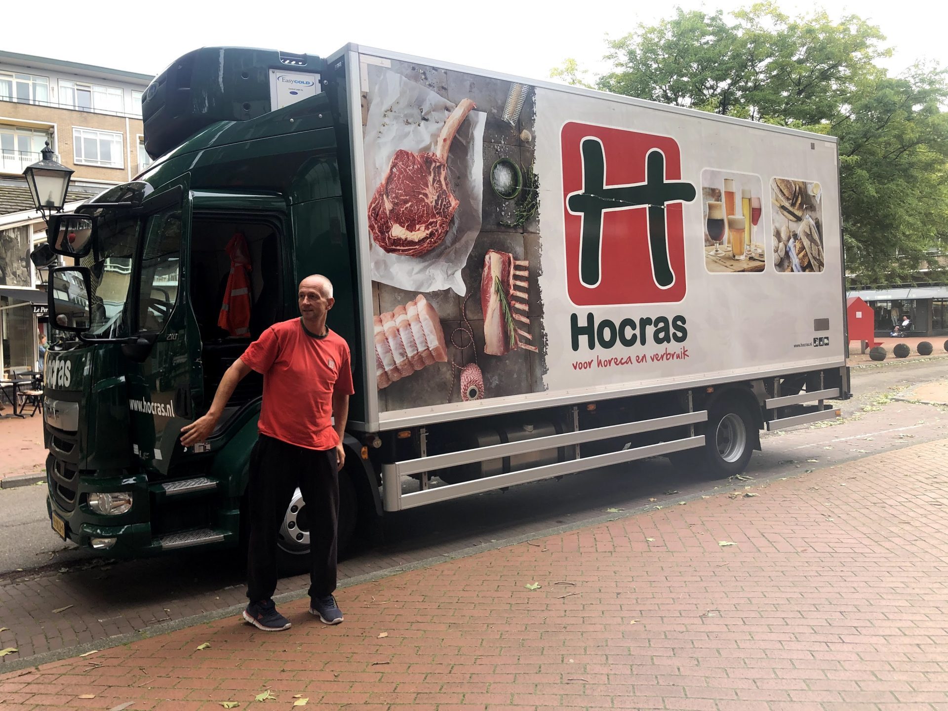 Een man in een rood shirt staat naast een groene vrachtwagen met Hocras-reclame op de zijkant.