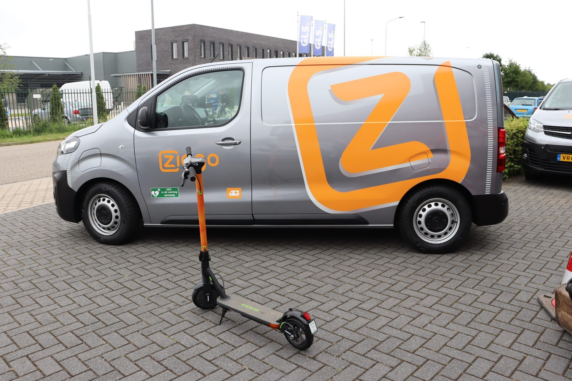 Grijze bestelbus van Ziggo met een oranje logoprint, naast een elektrische step op een parkeerplaats.