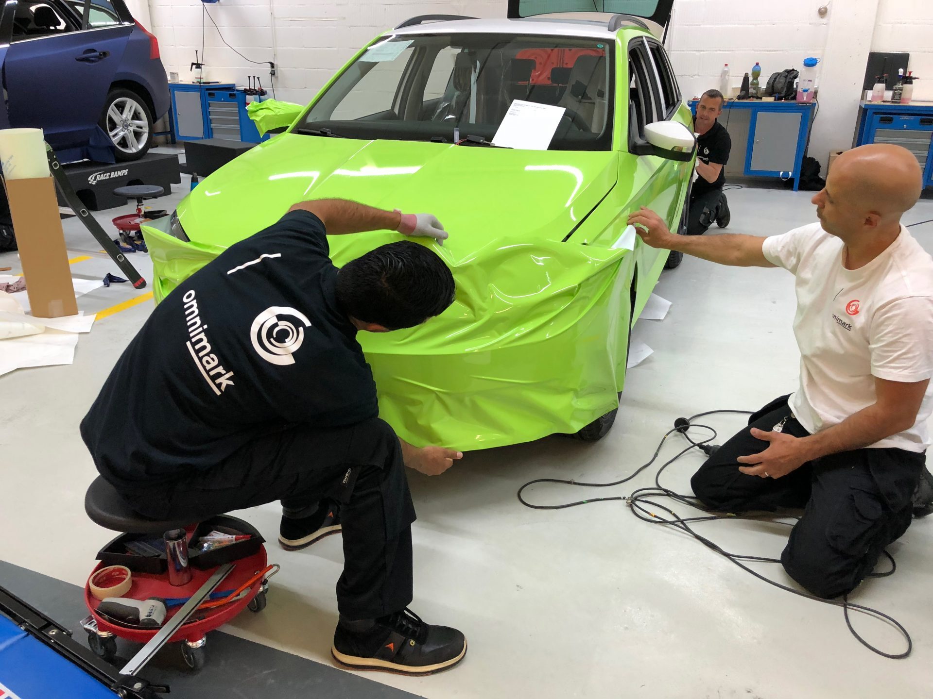 Twee mannen zijn bezig met het aanbrengen van een felgroene carwrap op een auto in een werkplaats.