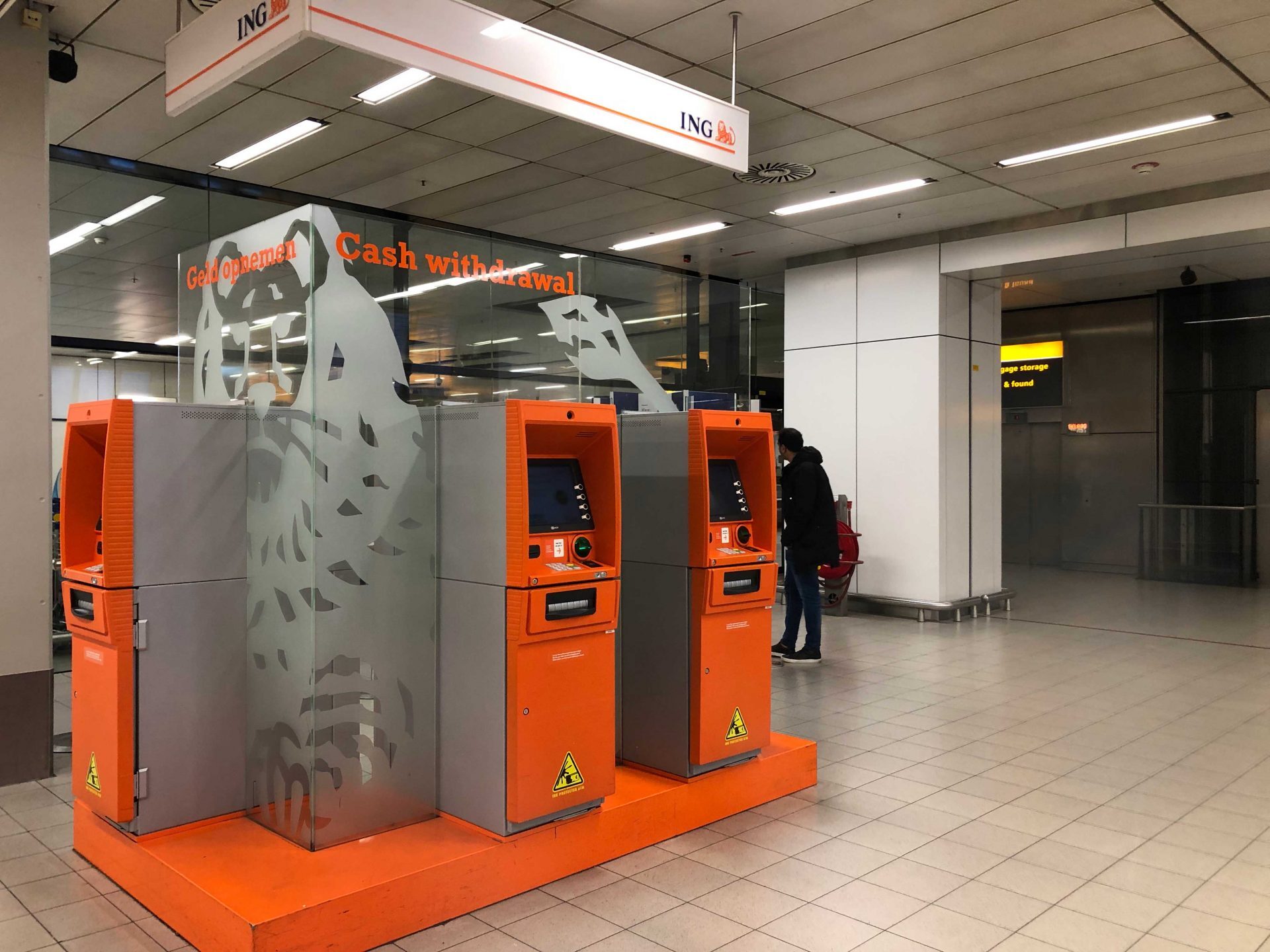 Twee oranje ING-geldautomaten staan opgesteld in een openbare ruimte met een mens in de buurt.