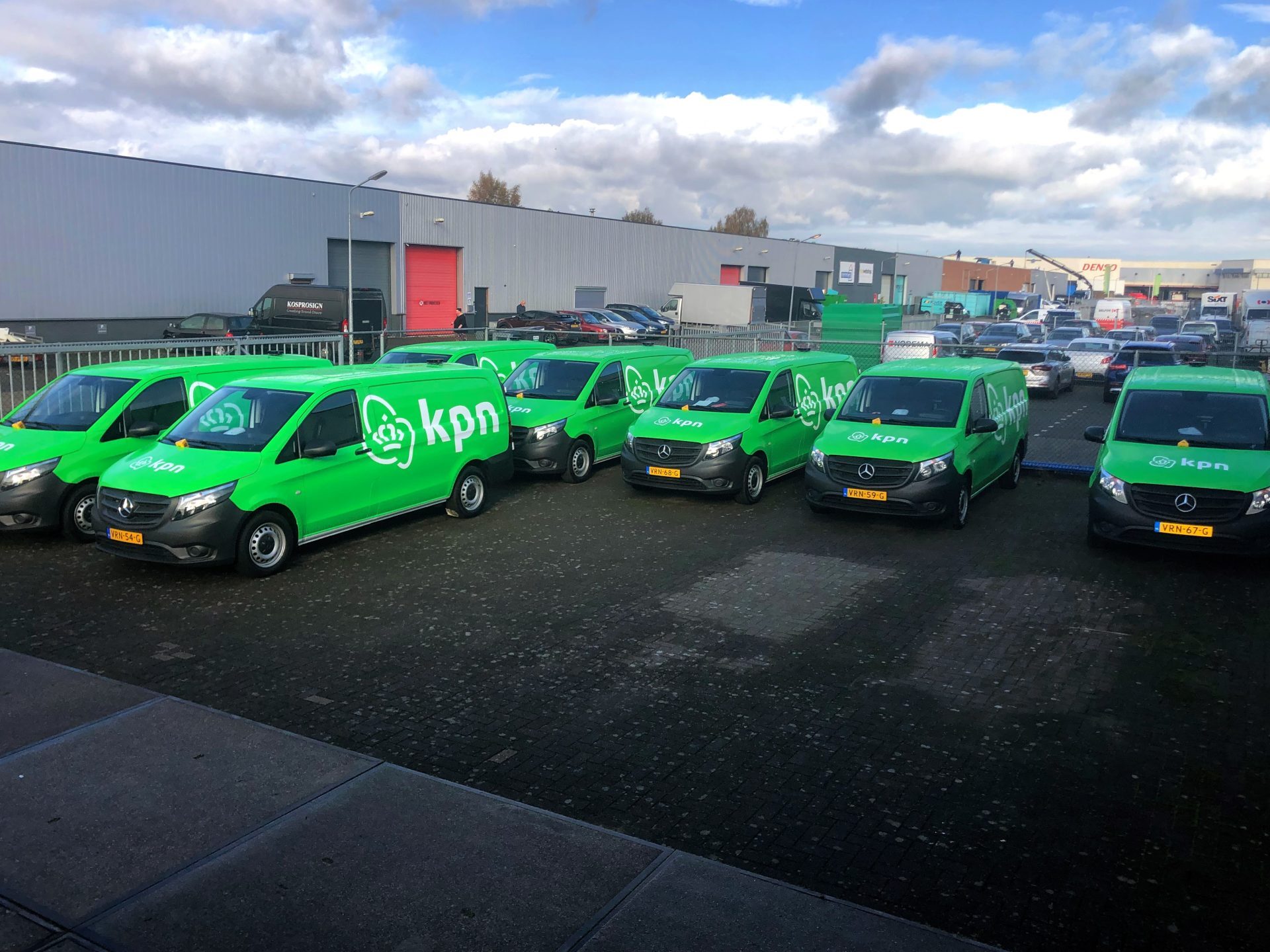 Een rij groene KPN-bestelwagens geparkeerd voor een industrieel gebouw op een parkeerterrein.