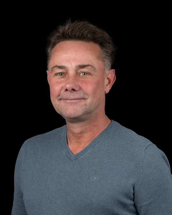 Portret van een glimlachende man in een blauwe trui met een zwarte achtergrond.