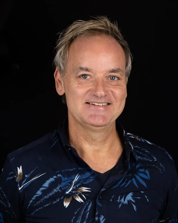 Een man in een donker overhemd met een patroon poseert lachend voor een zwarte achtergrond.