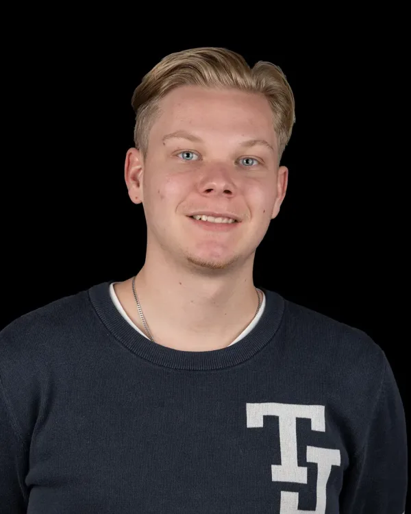 Een jongeman met blond haar, gekleed in een donkerblauwe trui, staat voor een zwarte achtergrond.