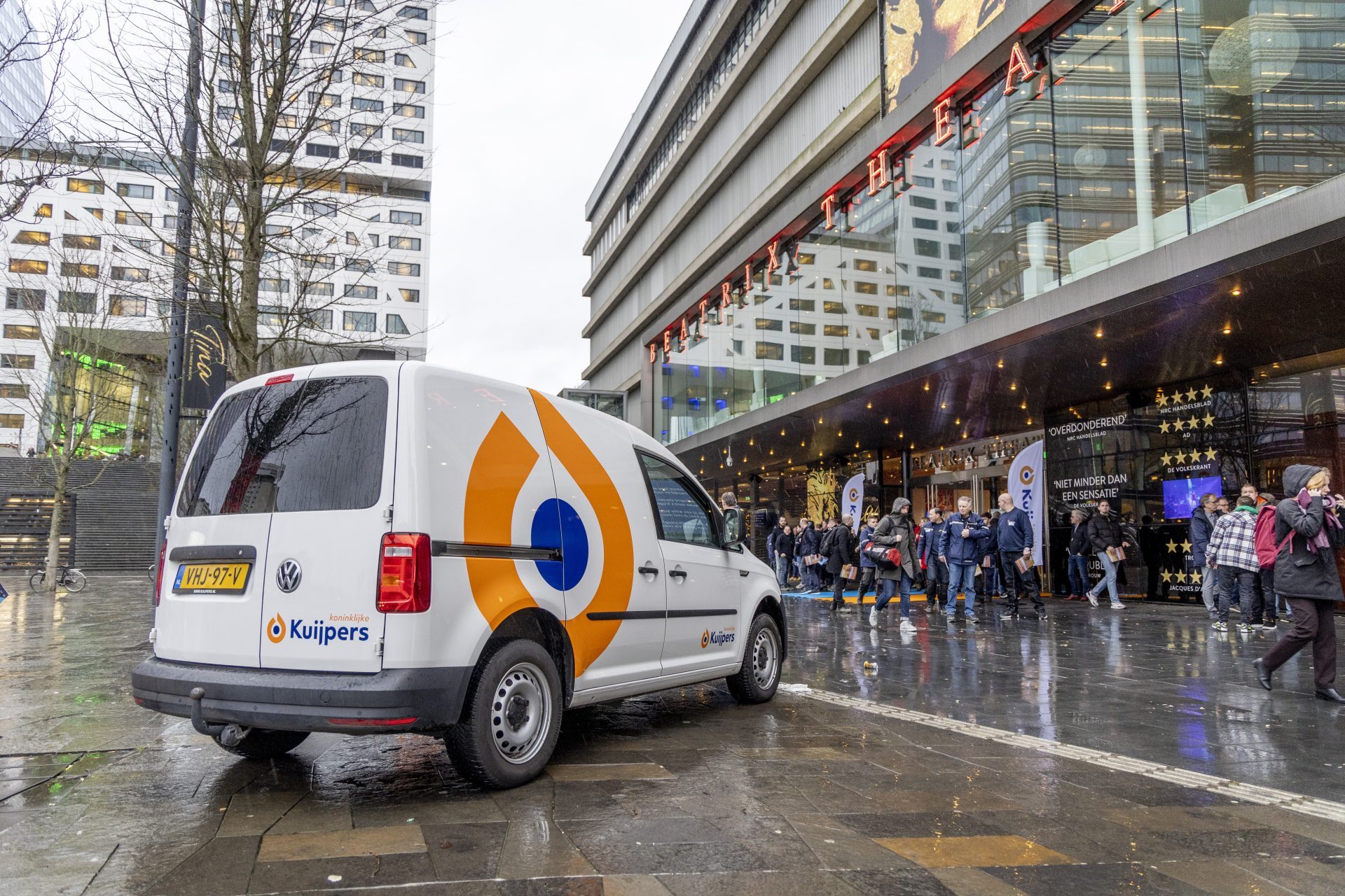 Een Volkswagen Caddy met het Kuijpers-logo staat geparkeerd voor het Beatrix Theater, waar mensen voor de ingang staan.