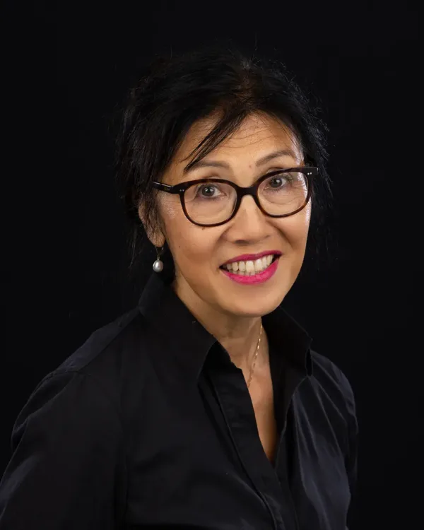 Portret van een glimlachende vrouw met bril tegen een zwarte achtergrond.