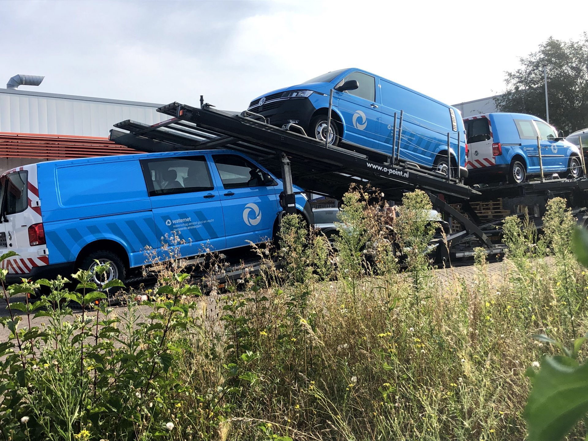 Blauwe Waternet-bestelwagens op een trailer, geparkeerd naast een gebouw.
