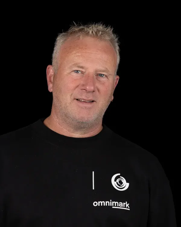 Een man met een zwart omnimark-shirt staat voor een donkere achtergrond.