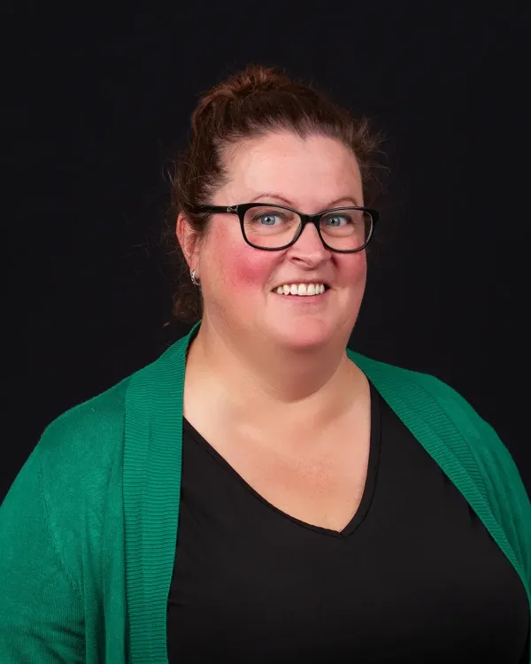 Een lachende vrouw met een groene trui en zwarte bril voor een donkere achtergrond.