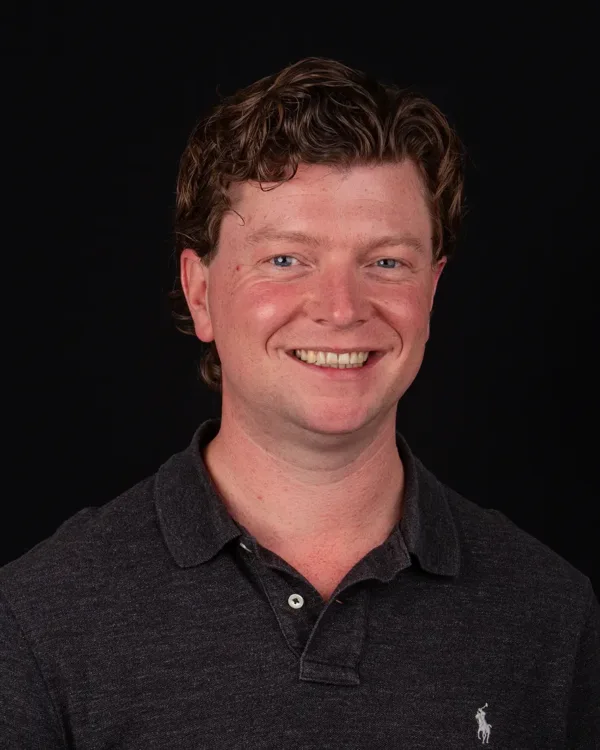 Portret van een lachende man in een donker poloshirt tegen een zwarte achtergrond.