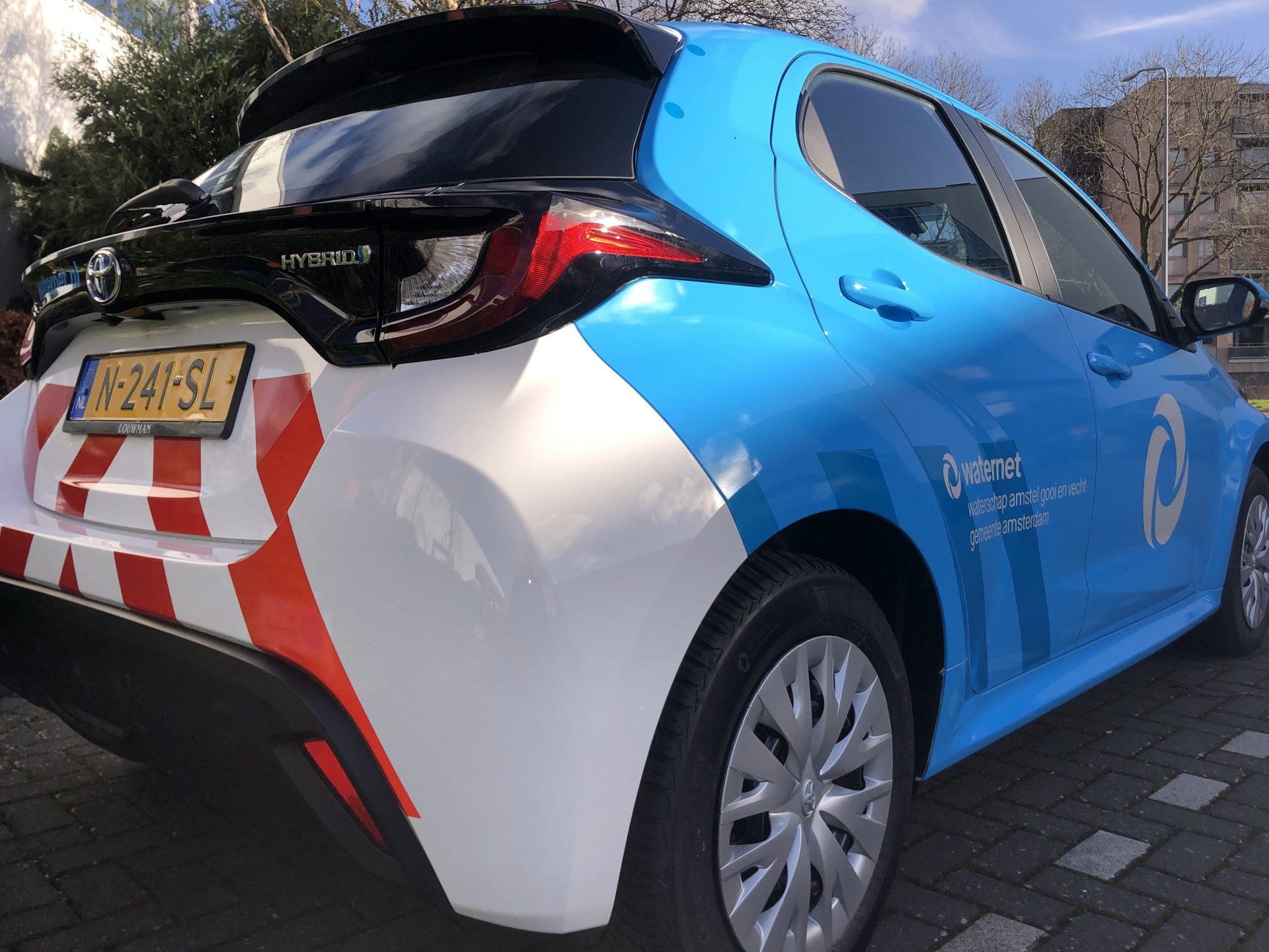 Een blauwe en witte Waternet hybride auto met kenteken N-241-SL, geparkeerd op een bestrate ondergrond.