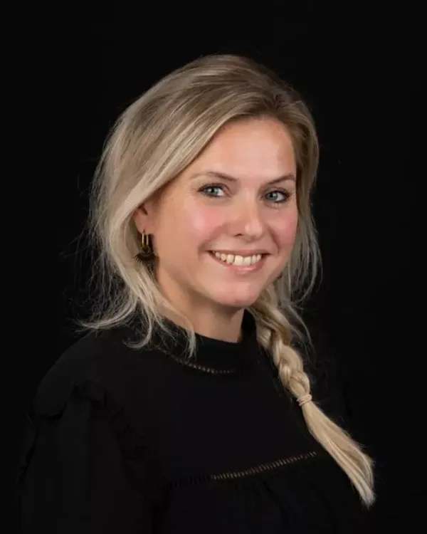 Portret van een glimlachende persoon met blond haar en een zwarte top tegen een zwarte achtergrond.