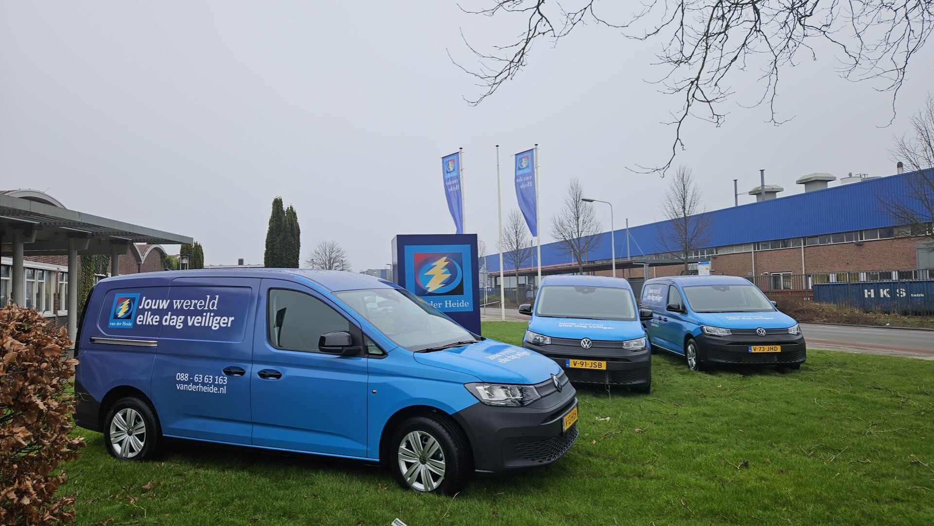 Drie blauwe bedrijfswagens met nieuwe huisstijl van Van der Heide geparkeerd voor hun gebouw.