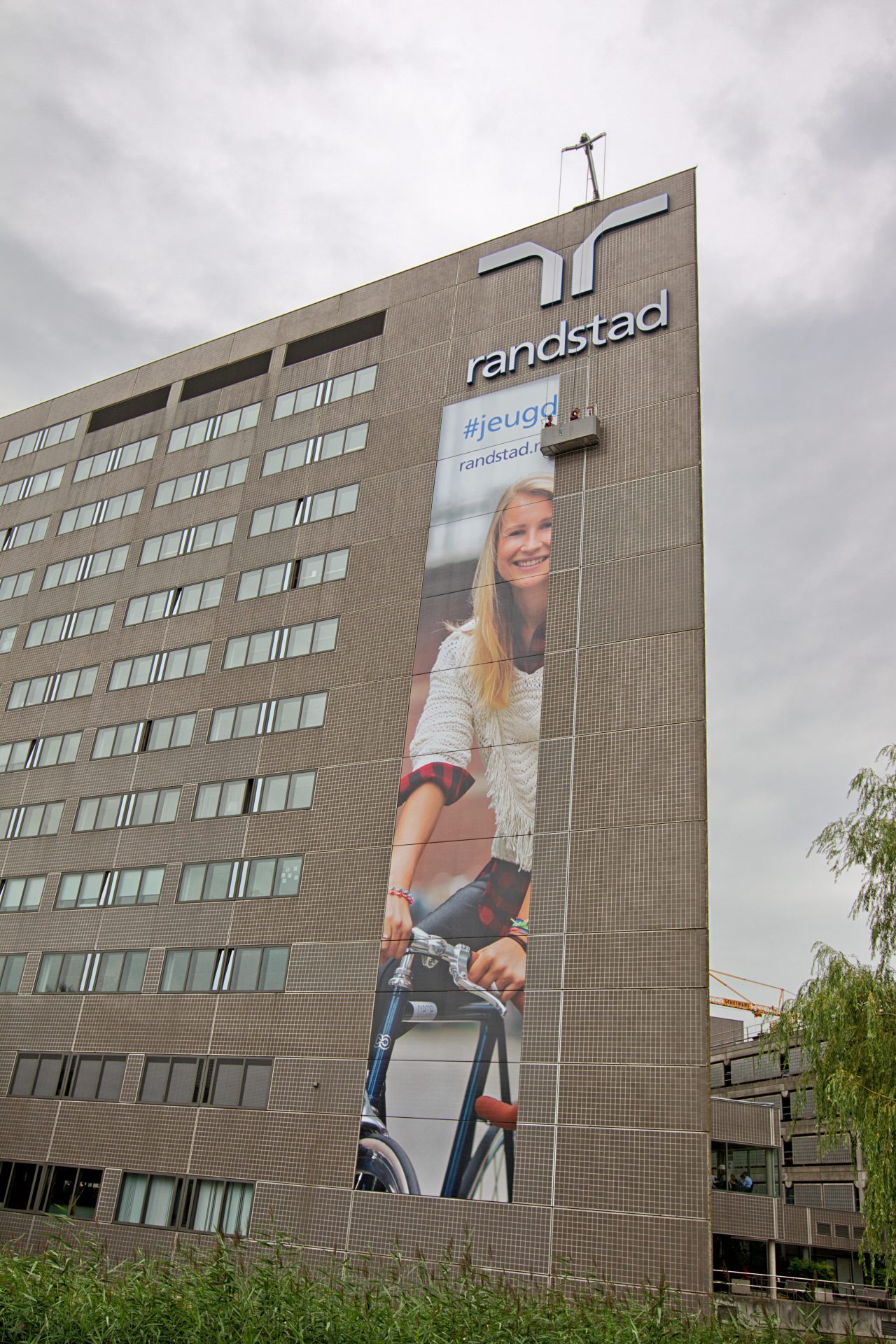 Hoog kantoorgebouw met groot Randstad-logo en een poster van een lachende vrouw op een fiets.
