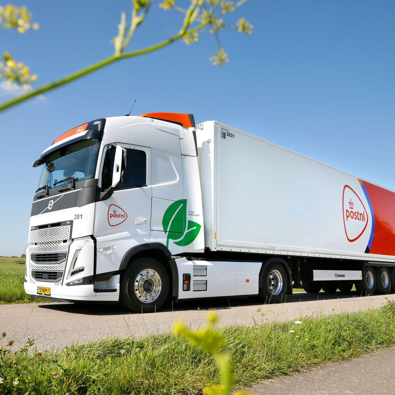 Een witte PostNL-vrachtwagen rijdt over een rustige weg omringd door groen met een blauwe lucht op de achtergrond.