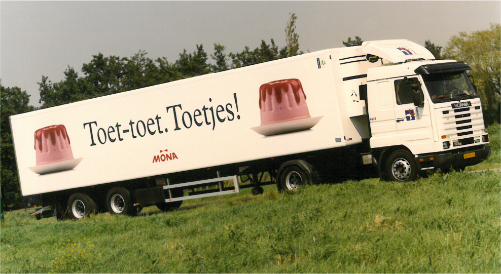 Een vrachtwagen met reclame voor Mona toetjes, geparkeerd in een groene omgeving.