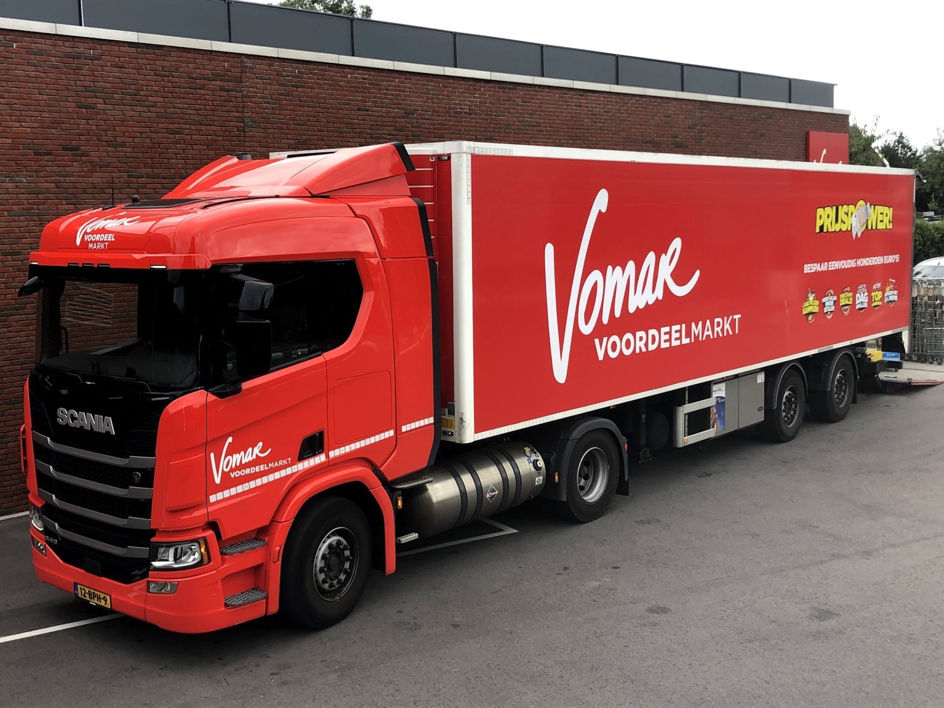 Een rode vrachtwagen van Vomar Voordeelmarkt geparkeerd voor een bakstenen muur.