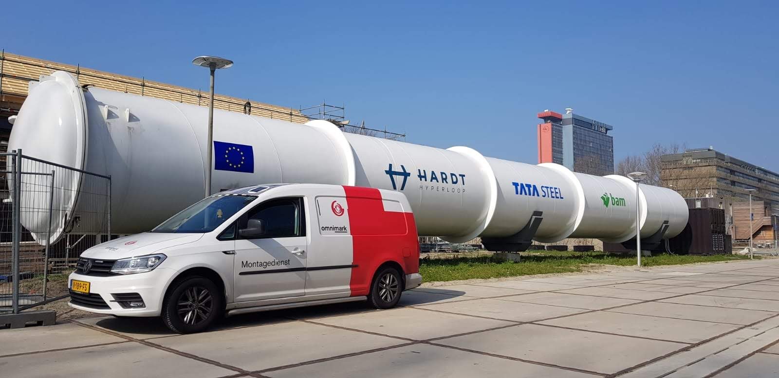 Een wagen van de Omnimark montagedienst staat geparkeerd voor een grote Hyperloop-testbuis met logo's van Hardt, Tata Steel en BAM.