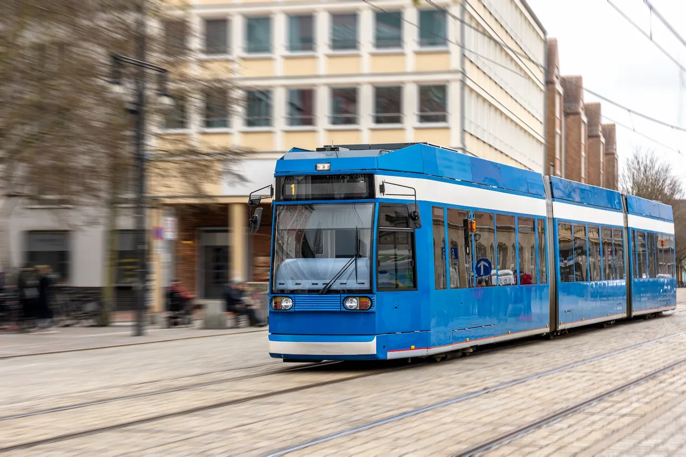 Blauwe tram rijdt op rails door een stedelijke straat met gebouwen op de achtergrond.