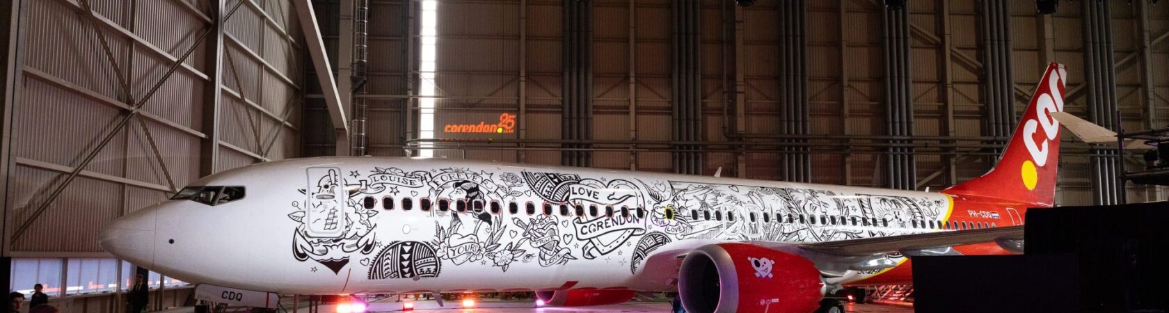 Een vliegtuig van Corendon met een unieke tattoo-achtige beschildering wordt gepresenteerd aan een publiek in een hangar.