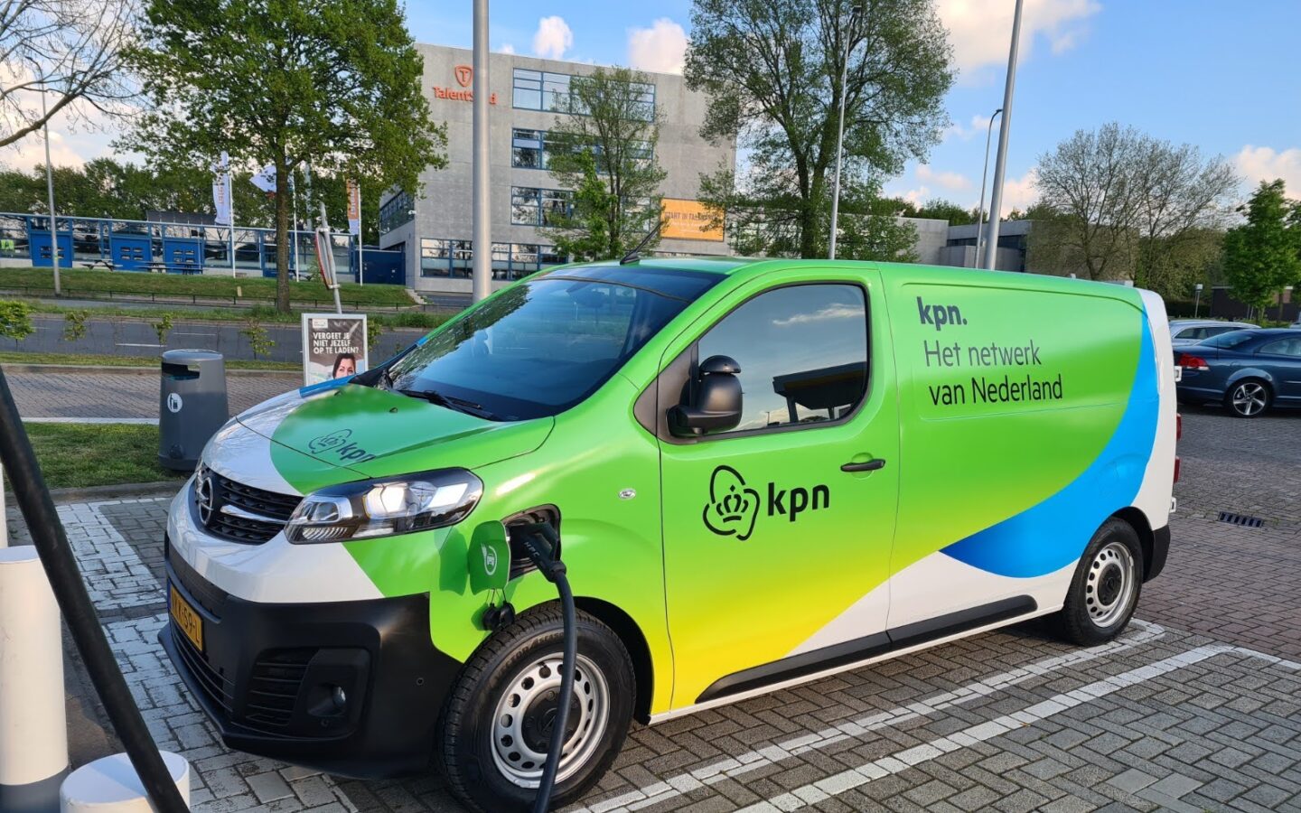 Groene en blauwe KPN-bestelwagen die aan het opladen is bij een laadstation.