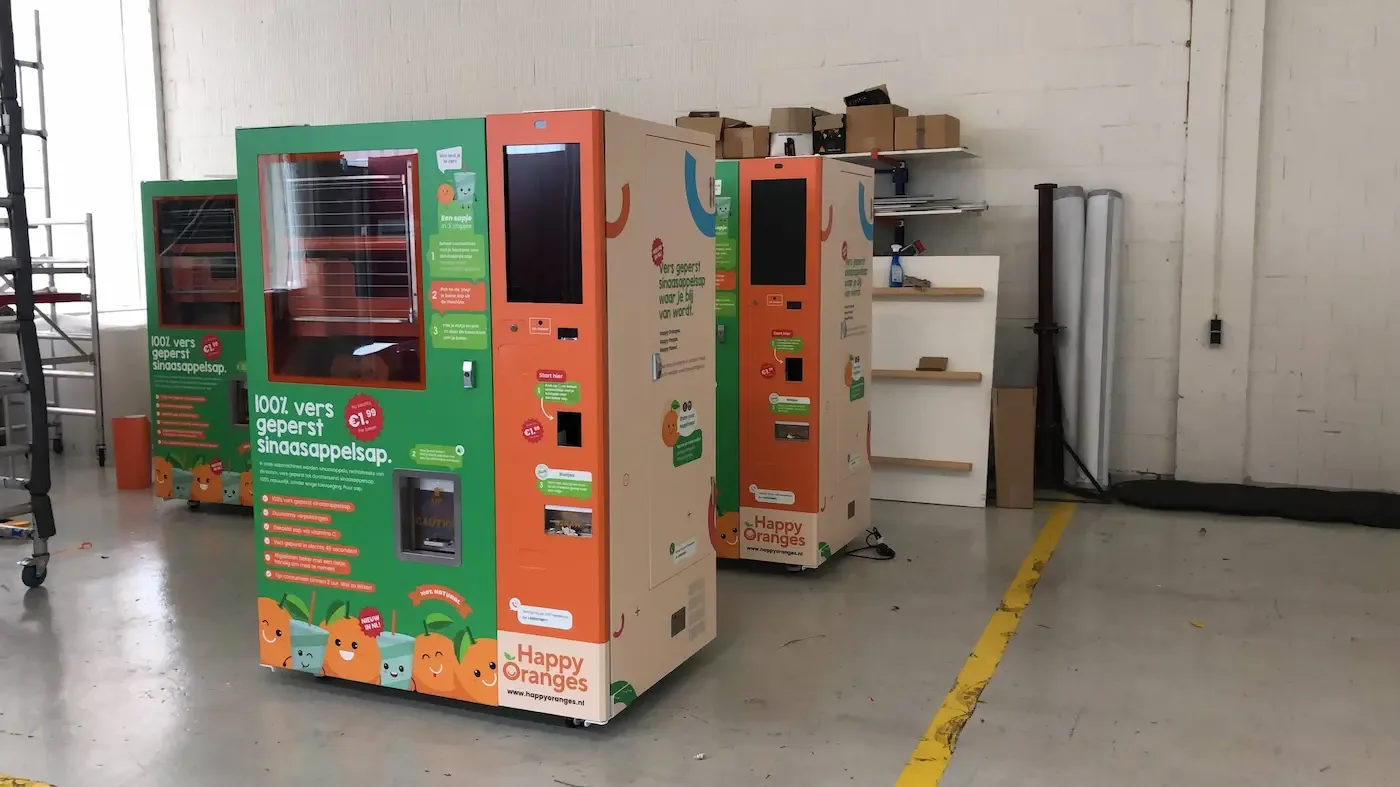 Twee sinaasappelsapautomaten van Happy Oranges staan in een magazijn met verschillende instructies en prijzen op de zijkant.