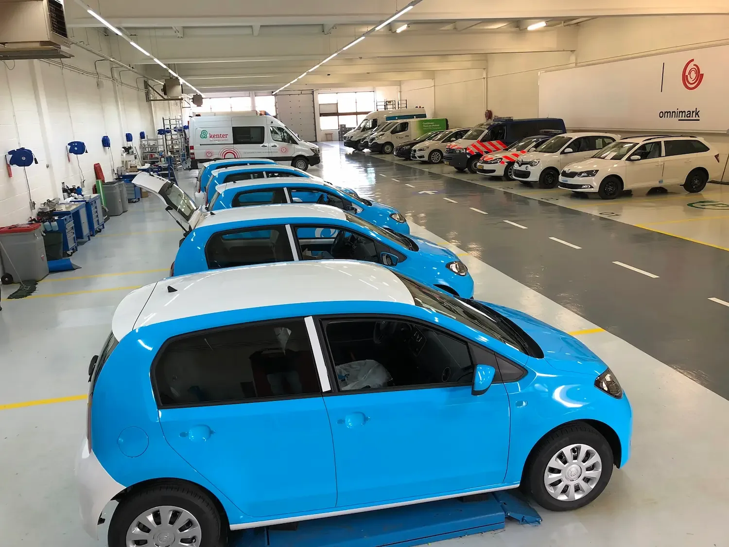 Een rij blauwe personenwagens in een werkplaats met meerdere bedrijfswagens op de achtergrond.