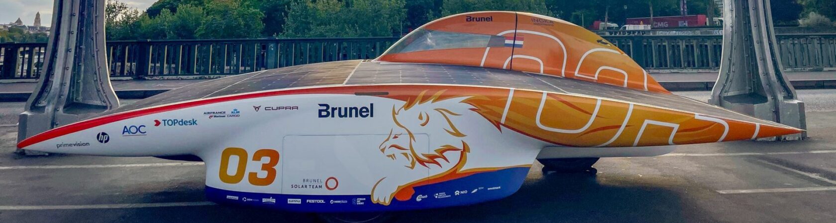 Een gestroomlijnde zonneauto met het nummer 03 en diverse sponsornamen, geparkeerd op een brug.
