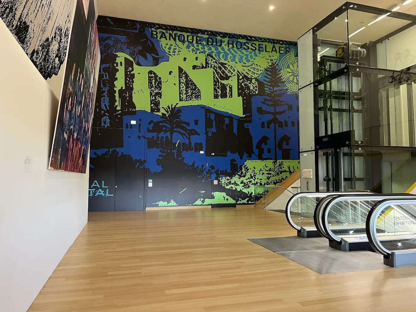 In een modern winkelcentrum is een kleurrijke muurschildering te zien naast een roltrap en een glazen lift.