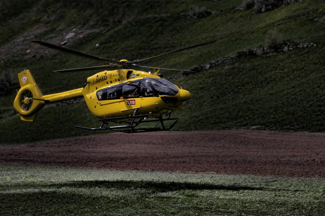 Een geel-zwarte helikopter zweeft boven een grasveld in een heuvelachtig landschap.