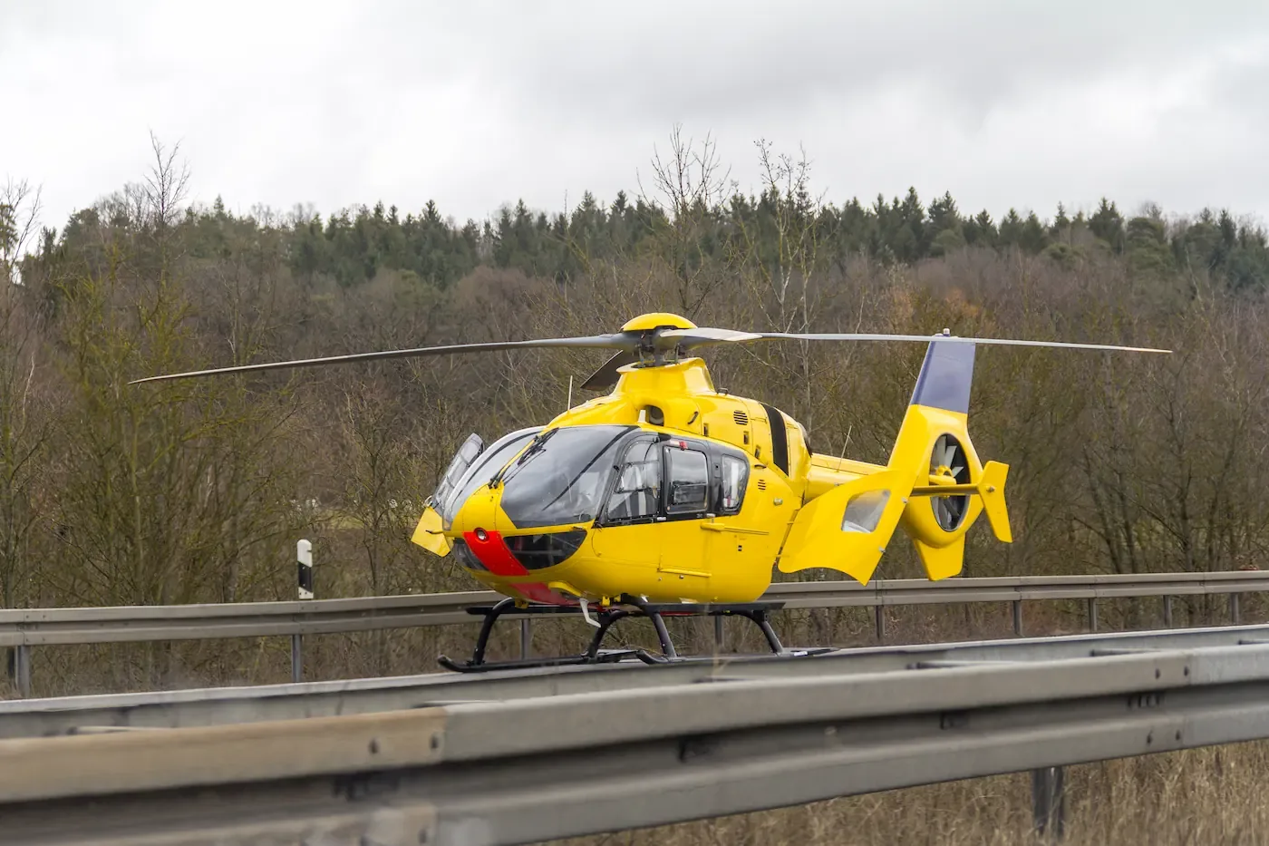 Een gele reddingshelikopter staat geparkeerd op een autoweg voor een bosrijke achtergrond.